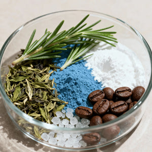 Glass bowl with various ingredients including herbs, coffee beans, and copper GHK blue powder on a light background